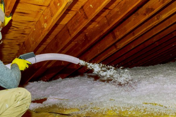 Technician spraying blown Fiberglass Insulation between Attic Trusses foam insulation construction foam from the gun to the roof Technician spraying blown Fiberglass Insulation between Attic Trusses foam insulation repair tool in the white protect suit applies a construction foam from the gun to the roof.